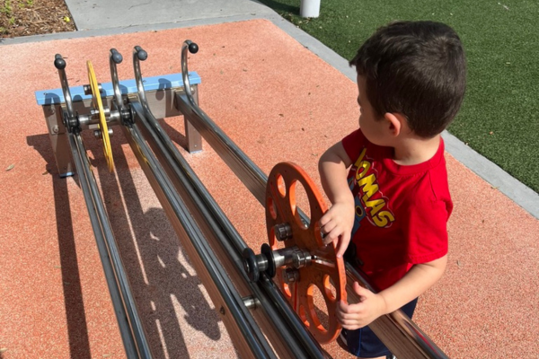 Image: A child with sensory needs is playing in a outdoor science exploration space at a museum. He is spinning a wheel down a metal track.