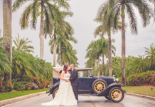 Miami Mom Collective Welcomes MIA Mom Sandra Velazquez Bride is dressed in a light cream wedding dressing being held by her husband to be in a black suit and black leather sneakers. They are leaning on a navy blue 1930s Model T car. They are surrounded by towering palm trees in Miami, FL.