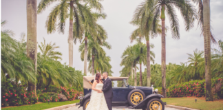 Bride is dressed in a light cream wedding dressing being held by her husband to be in a black suit and black leather sneakers. They are leaning on a navy blue 1930s Model T car. They are surrounded by towering palm trees in Miami, FL.