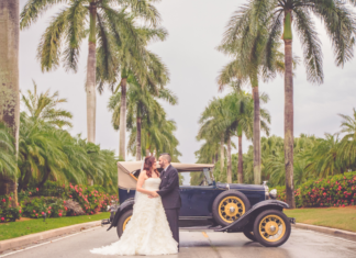 Bride is dressed in a light cream wedding dressing being held by her husband to be in a black suit and black leather sneakers. They are leaning on a navy blue 1930s Model T car. They are surrounded by towering palm trees in Miami, FL.