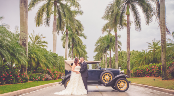 Bride is dressed in a light cream wedding dressing being held by her husband to be in a black suit and black leather sneakers. They are leaning on a navy blue 1930s Model T car. They are surrounded by towering palm trees in Miami, FL.