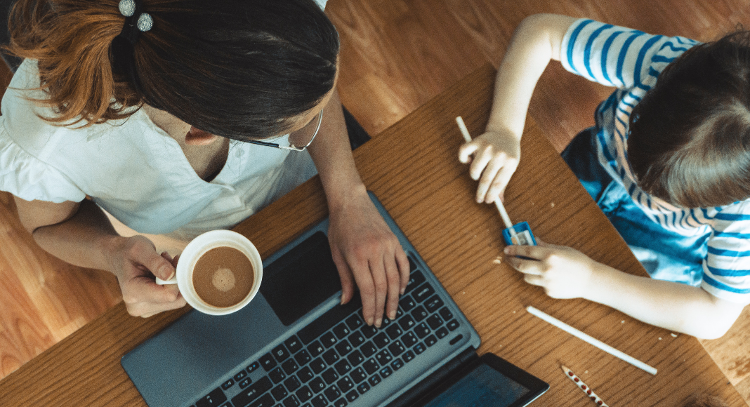 Image: A woman sits at a table next to a child, with a cup of coffee and a laptop