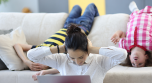 Image: A mom sits in front of a laptop with her hands on her ears, while two children hang upside down on the couch