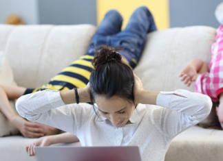 Image: A mom sits in front of a laptop with her hands on her ears, while two children hang upside down on the couch
