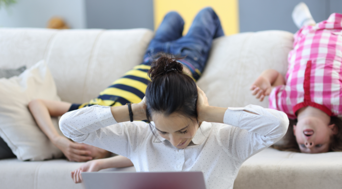 Image: A mom sits in front of a laptop with her hands on her ears, while two children hang upside down on the couch