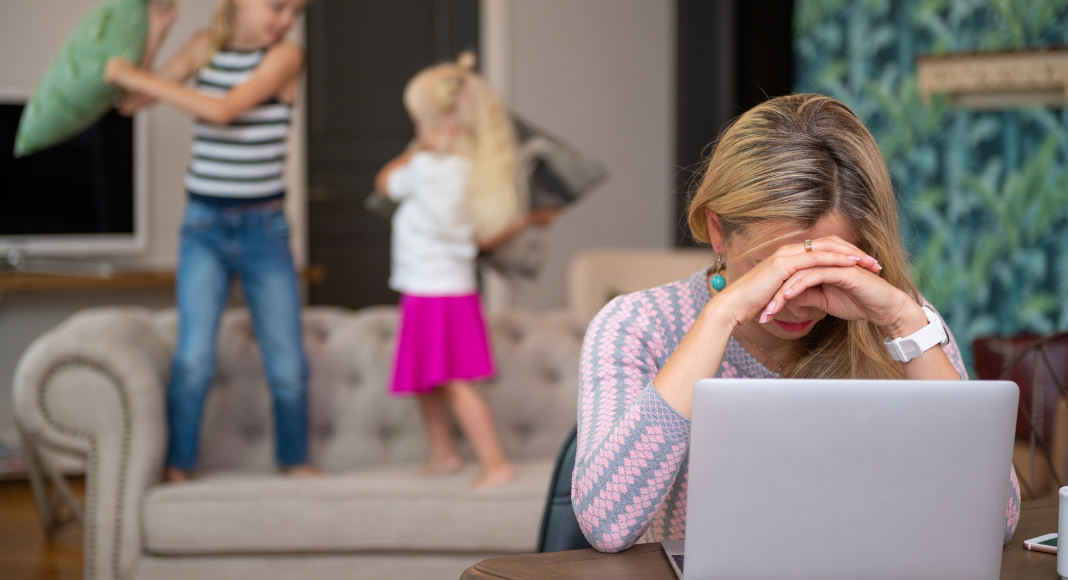 Image: A mom struggles to work while her kids have a pillow fight on the couch behind her