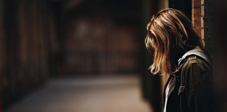 Image: A woman stands with her back to a wall, looking down at the ground