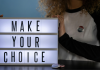 Image: A woman with an "I voted" sticker holds a letterboard that reads, "Make Your Choice"