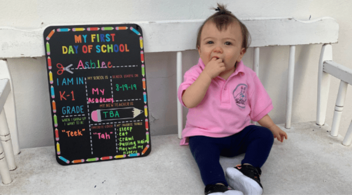 A toddler sits next to a My First Day of School board