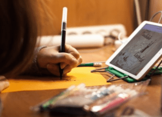 A child sitting in front of a tablet at a table, writing with a marker