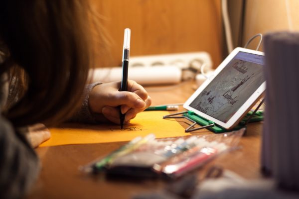 A child sitting in front of a tablet at a table, writing with a marker