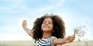 A young girl stands in a field with her arms spread wide, eyes closed and smiling. She is holding a bunch of wildflowers and the wind is blowing her hair.