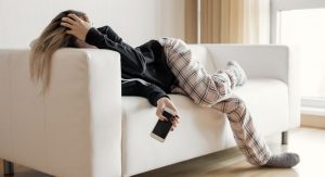 Image: A burned out woman lies on the couch, holding her head in one hand and her phone in the other