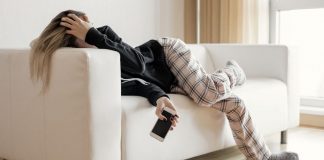 A burned out woman lies on the couch, holding her head in one hand and her phone in the other