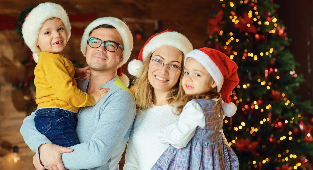 Image; A family wearing Santa hats and standing in front of a Christmas tree (Healthy Holidays: A Survival Guide | Part 2 Dina Garcia Contributor Miami Mom Collective)