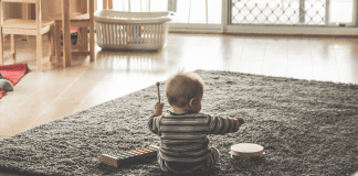 A baby playing on a grey rug in a minimalist room. The baby is playing with musical toys.