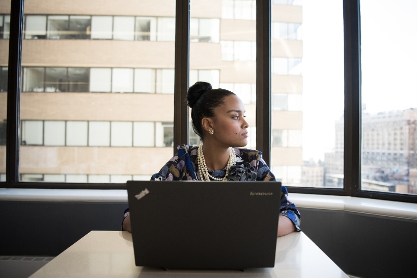Image: A woman sits in a corporate office with a laptop, looking out a window