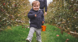Image: a little boy walks in front of his mother towards the camera in an apple orchard. He is smiling, carrying a bucket, and wearing a jacket, sweatpants, and Adidas sneakers. (Toddler Shoes: What to Look for Brittany Aquart Contributor Miami Mom Collective)