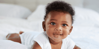 Image description: a baby smiling and wearing a white onesie while doing tummy time on a bed. (Tummy Time Without Tears: Tips From a Pediatric Physical Therapist Brittany Aquart Contributor Miami Mom Collective)