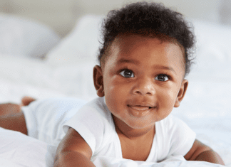 Tummy Time Without Tears: Tips From a Pediatric Physical Therapist Image description: a baby smiling and wearing a white onesie while doing tummy time on a bed. (Tummy Time Without Tears: Tips From a Pediatric Physical Therapist Brittany Aquart Contributor Miami Mom Collective)