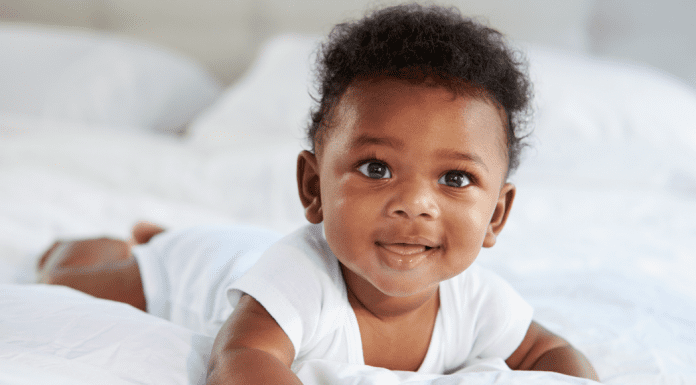 Image description: a baby smiling and wearing a white onesie while doing tummy time on a bed. (Tummy Time Without Tears: Tips From a Pediatric Physical Therapist Brittany Aquart Contributor Miami Mom Collective)