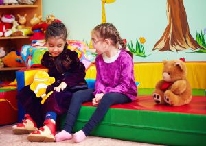 Image Description: Two girls are sitting together in a brightly colored room. One girl is wearing adaptive shoes and appears to have a physical disability.