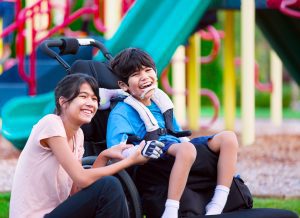 Image Description: A boy and a girl are in front of a playground. The girl is able-bodied and the boy is sitting in a wheelchair.