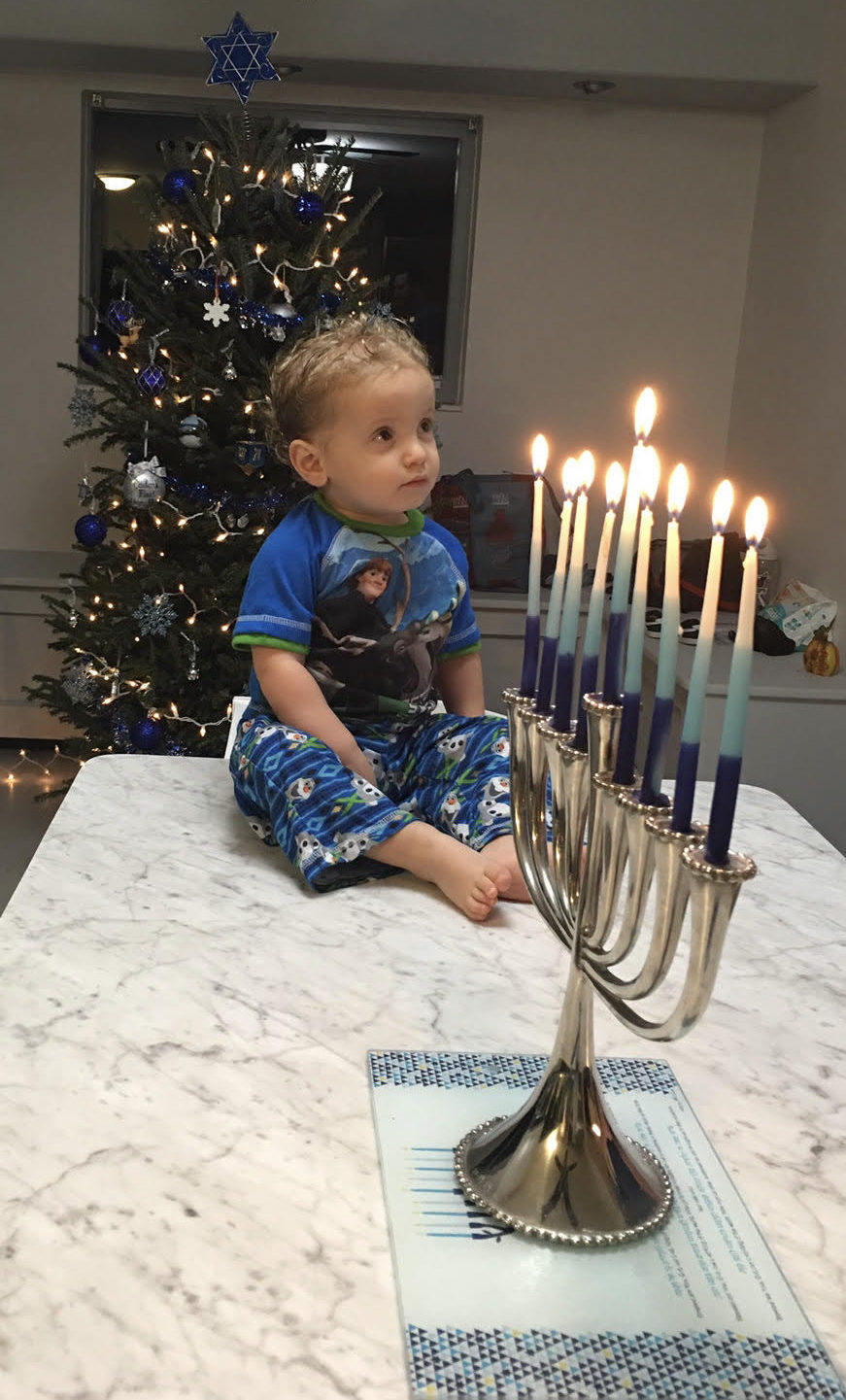 Image: A child sits next to a lit menorah, and with a holiday tree decorated for Hanukkah in the background