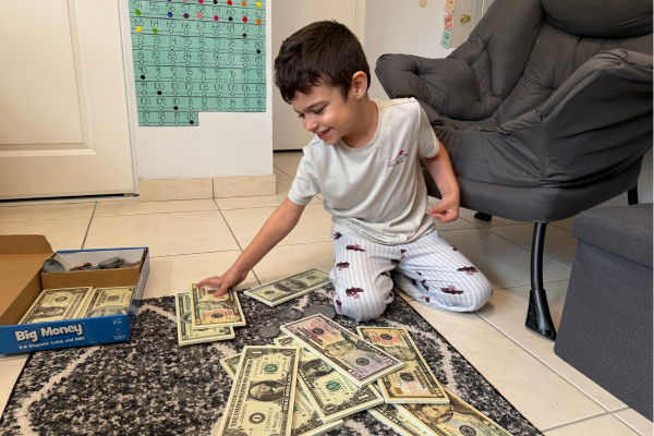 A homeschool neurodivergent child is smiling as he sits on the floor. He is choosing from a stack of large play money.