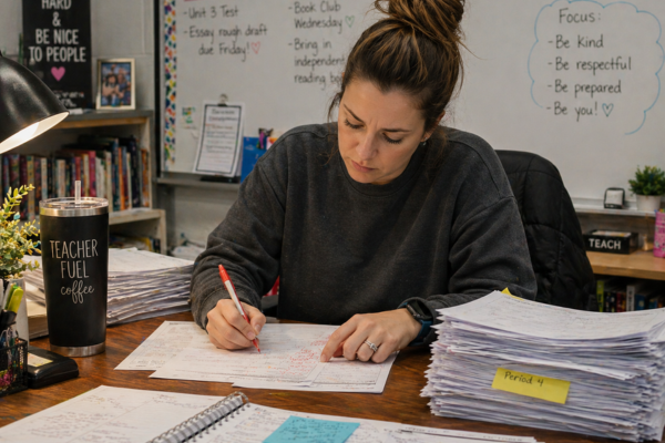 A tired teacher is grading a large stack of papers on her desk. 