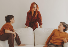 Image: A mother stands behind a couch where her two children are seated