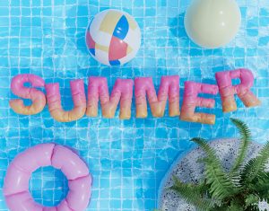 Image: Inflatable letters spelling out SUMMER float on the surface of a pool, framed by beach balls and an inner tube
