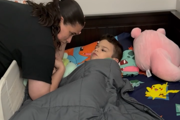 A mom is putting her son to sleep. She is leaning over the side of the bed as he looks at her while laying down.