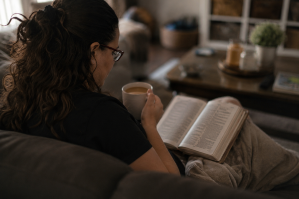 A woman is sitting on the couch holding a coffee cup in hand and the Bible sitting open on her lap.