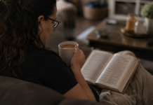 Image: A woman sits on a couch with a Bible open on her lap and a cup of coffee in her hand