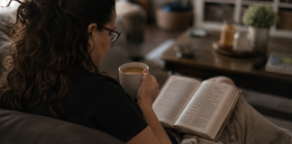 Holding on to Faith as a Full-Time Caregiver Image: A woman sits on a couch with a Bible open on her lap and a cup of coffee in her hand