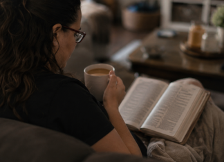 Image: A woman sits on a couch with a Bible open on her lap and a cup of coffee in her hand