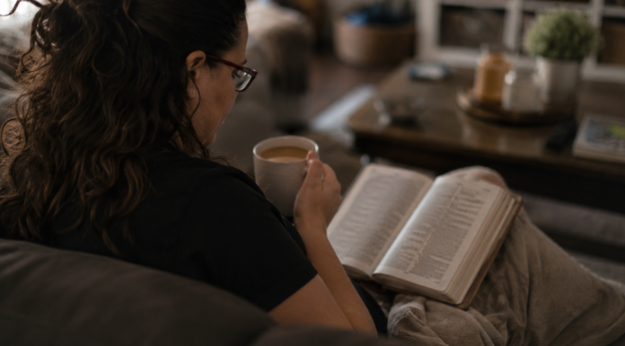 Holding on to Faith as a Full-Time Caregiver Image: A woman sits on a couch with a Bible open on her lap and a cup of coffee in her hand