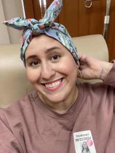 Image: A woman wearing a scarf smiles from a chemo treatment chair