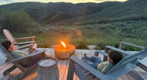 Image: Two children sit in Adirondack chairs around a campfire, with a view of mountains in the background