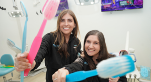 Image: Dr. Smith and Dr. Lenard of All Smiles Pediatric Dentistry smile for the camera, each holding a large colorful toothbrush