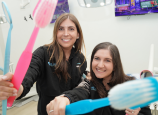 All Smiles Pediatric Dentistry: Comfort, Care & Community Image: Dr. Smith and Dr. Lenard of All Smiles Pediatric Dentistry smile for the camera, each holding a large colorful toothbrush