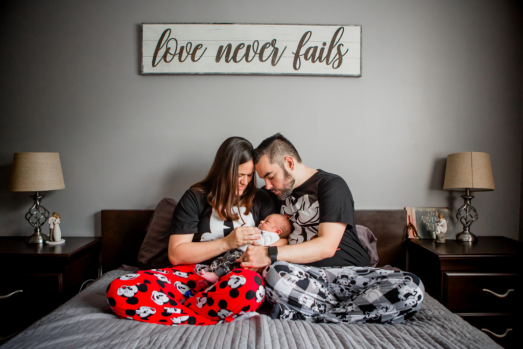 Mother and father sitting together on a bed, lovingly holding their newborn baby beneath a sign that reads “love never fails.”