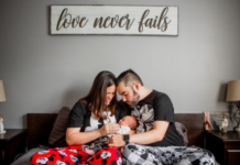 Grief Doesn’t Stop the Rest of Life from Moving Mother and father sitting together on a bed, lovingly holding their newborn baby beneath a sign that reads “love never fails.”