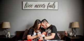 Grief Doesn’t Stop the Rest of Life from Moving Mother and father sitting together on a bed, lovingly holding their newborn baby beneath a sign that reads “love never fails.”
