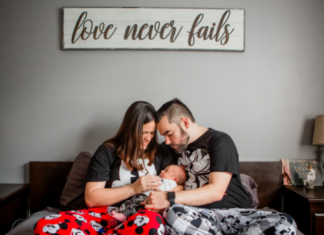 Grief Doesn’t Stop the Rest of Life from Moving Mother and father sitting together on a bed, lovingly holding their newborn baby beneath a sign that reads “love never fails.”