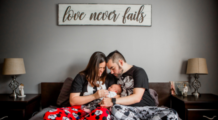 Grief Doesn’t Stop the Rest of Life from Moving Mother and father sitting together on a bed, lovingly holding their newborn baby beneath a sign that reads “love never fails.”